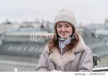 Young woman wears warm attire with winter hat while standing on rooftop in city during overcast day Young woman wears warm attire with winter hat while standing on rooftop in city during overcast day 129915197