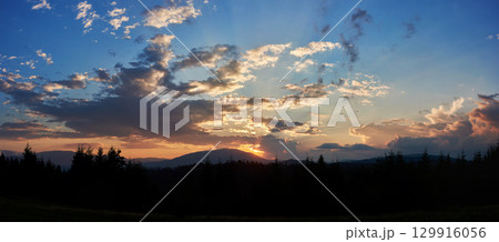 Serene evening sky with hues of blue and soft pink clouds as sun sets. Horizon glows gently, casting light over silhouetted pine trees and rolling hills, creating peaceful and expansive landscape. 129916056