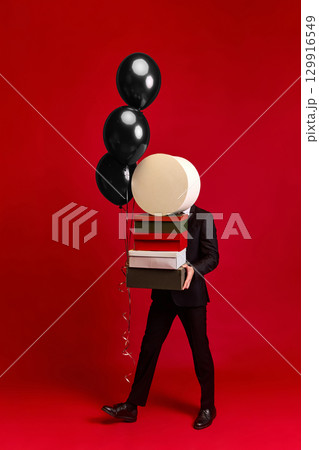 Person walking with gift boxes and black balloons on vivid red background Person walking with gift boxes and black balloons on vivid red background 129916549