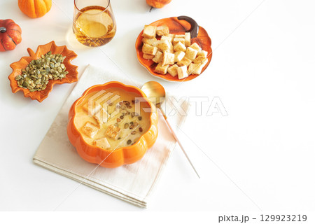 Creamy pumpkin soup with croutons and seeds, served in a pumpkin-shaped bowl on white wooden table. Top view Creamy pumpkin soup with croutons and seeds, served in a pumpkin-shaped bowl on white wooden table. Top view 129923219