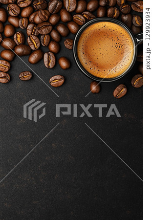 A cup of coffee sits on a dark, textured background, surrounded by scattered roasted coffee beans. The image is shot from a top-down perspective. 129923934