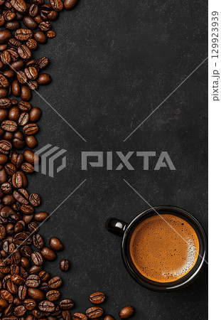 A cup of coffee sits on a dark, textured background, surrounded by scattered roasted coffee beans. The image is shot from a top-down perspective. A cup of coffee sits on a dark, textured background, surrounded by scattered roasted coffee beans. The image is shot from a top-down perspective. 129923939
