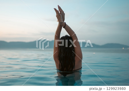 beautiful woman swimming in luxury pool at sunset flowing into sea, elegant vacation and tropical serenity, stunning girl relaxing in lavish resort pool at sunrise 129923980