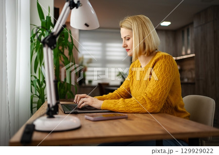 Focused woman in yellow sweater working on laptop at home desk with bright lamp and plants in a cozy atmosphere Focused woman in yellow sweater working on laptop at home desk with bright lamp and plants in a cozy atmosphere 129929022