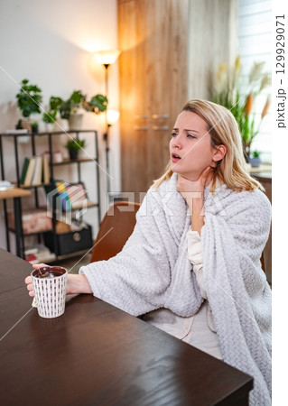 Woman in cozy blanket holds cup while feeling discomfort at home Woman in cozy blanket holds cup while feeling discomfort at home 129929071