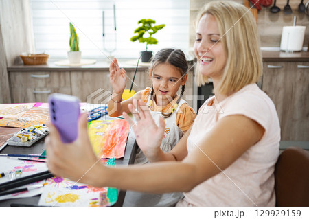 Mother and daughter engaged in a cheerful video call while painting together at home in a cozy kitchen setting Mother and daughter engaged in a cheerful video call while painting together at home in a cozy kitchen setting 129929159