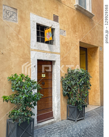 ROME, ITALY - July 30, 2025 Narrow street view showing an old yellow building facade with a wooden door and a colorful flag hangs above. Two potted green plants flank the entrance ROME, ITALY - July 30, 2025 Narrow street view showing an old yellow building facade with a wooden door and a colorful flag hangs above. Two potted green plants flank the entrance 129929368