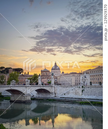 Scenic sunset over the Tiber River in Rome. The Ponte Sant'Angelo visible in the foreground with Saint Peter's Basilica in background. The colorful sky and city buildings reflects in the calm waters 129929370