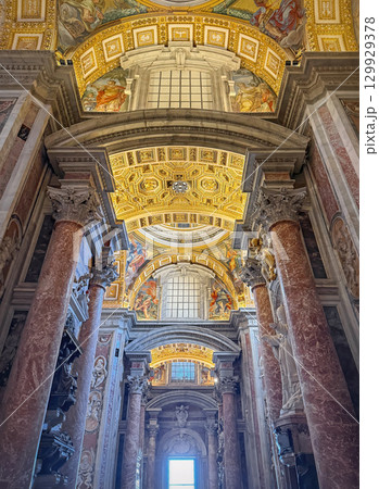 A view looking up inside the majestic St. Peter's Basilica. Massive reddish marble columns frame the ornate ceiling, richly decorated with gilded patterns and detailed religious frescoes A view looking up inside the majestic St. Peter's Basilica. Massive reddish marble columns frame the ornate ceiling, richly decorated with gilded patterns and detailed religious frescoes 129929378