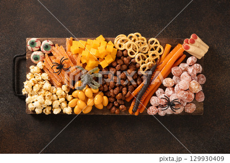 Halloween sweet charcuterie board with pumpkin, popcorn, dried fruits, color chocolate candy, spider, crackers on dark brown background. Festive food for kids party. View from above. 129930409