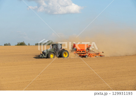A tractor towing a seeder creates dust clouds while performing field work 129931193