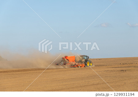 A tractor towing a seeder creates dust clouds while performing field work 129931194