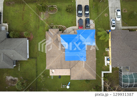 Top view of leaking house roof covered with protective tarp sheets against rain water leaks until replacement of asphalt shingles. Damage of building rooftop as aftermath of hurricane Ian in Florida Top view of leaking house roof covered with protective tarp sheets against rain water leaks until replacement of asphalt shingles. Damage of building rooftop as aftermath of hurricane Ian in Florida 129931387