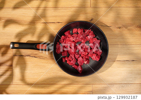 Fresh diced beef cooking in a skillet on a wooden surface with natural light streaming in 129932187