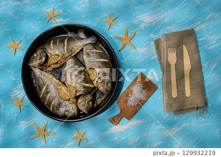 Four dorado fish are arranged in a black dish with lemon slices resting on a vibrant blue background Four dorado fish are arranged in a black dish with lemon slices resting on a vibrant blue background 129932219