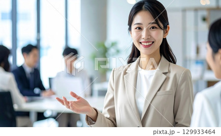 ベージュスーツの日本人女性がオープンオフィスで同僚と会話する笑顔ポートレート 129933464