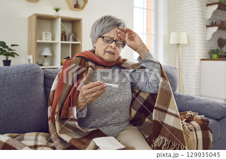 Unhappy sick senior woman being treated at home measures body temperature with thermometer. 129935045