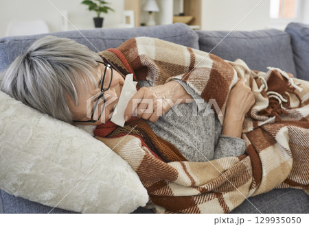 Senior sick woman lying on sofa at home wrapped in a blanket suffering from runny nose. Senior sick woman lying on sofa at home wrapped in a blanket suffering from runny nose. 129935050