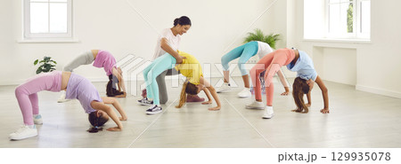 Woman trainer helps flexible children to do bridge exercise at gymnastics studio Woman trainer helps flexible children to do bridge exercise at gymnastics studio 129935078