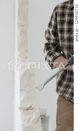 Close up of unrecognizable male construction worker wearing beige checkered shirt and protective gloves, is demolishing white wall with rotary hammer drill, generating dust. Renovation concept Close up of unrecognizable male construction worker wearing beige checkered shirt and protective gloves, is demolishing white wall with rotary hammer drill, generating dust. Renovation concept 129938718