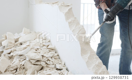Unknown male construction worker wearing red checkered shirt jeans and protective gloves, is demolishing white wall with rotary hammer drill, generating debris on the floor, close up view. Renovation 129938731