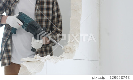 Close up of unrecognizable male construction worker wearing beige checkered shirt and protective gloves, is demolishing white wall with rotary hammer drill, generating dust, close up view. Renovation 129938748
