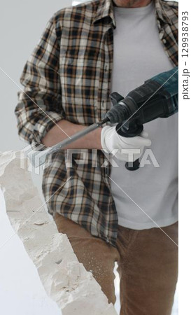 Close up of unknown male construction worker wearing beige checkered shirt and protective gloves, is demolishing white wall with rotary hammer drill, generating dust. Renovation project Close up of unknown male construction worker wearing beige checkered shirt and protective gloves, is demolishing white wall with rotary hammer drill, generating dust. Renovation project 129938793