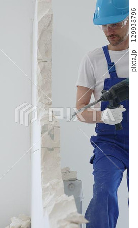 Male construction worker wearing blue overalls, protective helmet and white gloves, is demolishing white wall with rotary hammer drill, generating dust, close up horizontal view 129938796
