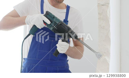 Unrecognizable male construction worker wearing blue overalls and protective white gloves, is demolishing white wall with rotary hammer drill, generating dust, close up horizontal view. Renovation 129938800