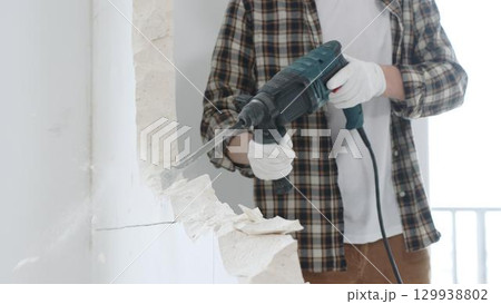 Close up of unrecognizable male construction worker wearing beige checkered shirt and protective gloves, is demolishing white wall with rotary hammer drill, generating dust, close up view. Renovation 129938802