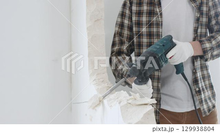 Close up of unrecognizable male construction worker wearing beige checkered shirt and protective gloves, is demolishing white wall with rotary hammer drill, generating dust, close up view. Renovation 129938804