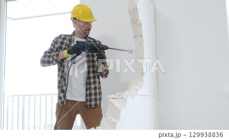 Male construction worker wearing beige checkered shirt, yellow hard hat and protective gloves, is demolishing white wall with rotary hammer drill, generating dust, medium long shot. Renovation project 129938836