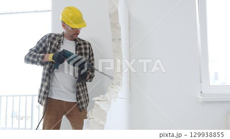 Man construction worker wearing beige checkered shirt , yellow hard hat and protective gloves, is demolishing white wall with rotary hammer drill, generating dust, medium long shot. Renovation concept 129938855