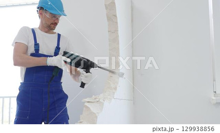 Man construction worker wearing blue overalls, hard hat and protective white gloves, is demolishing white wall with rotary hammer drill, generating dust, close up horizontal view. Renovation concept Man construction worker wearing blue overalls, hard hat and protective white gloves, is demolishing white wall with rotary hammer drill, generating dust, close up horizontal view. Renovation concept 129938856