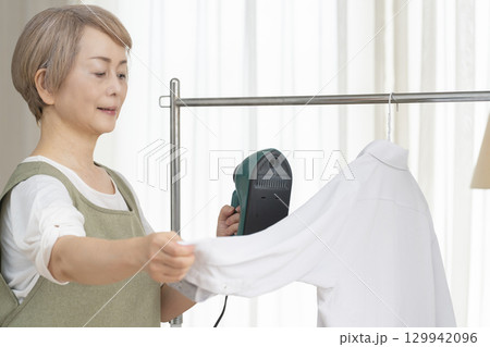 リビングでハンガーに掛けた衣類にスチームアイロンをかけるシニア   129942096