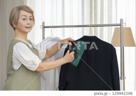 リビングでハンガーに掛けた衣類にスチームアイロンをかけるシニア   129942106