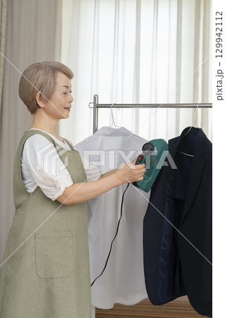 リビングでハンガーに掛けた衣類にスチームアイロンをかけるシニア   129942112