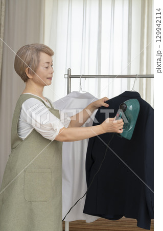 リビングでハンガーに掛けた衣類にスチームアイロンをかけるシニア   129942114
