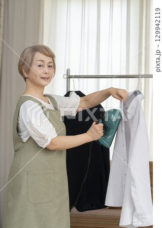 リビングでハンガーに掛けた衣類にスチームアイロンをかけるシニア   129942119