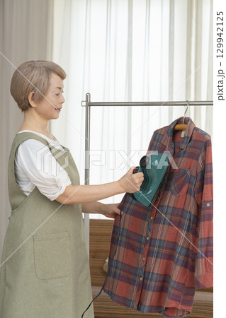リビングでハンガーに掛けた衣類にスチームアイロンをかけるシニア   129942125