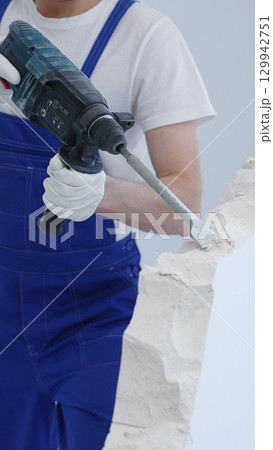 Construction worker demolishing white wall with rotary hammer drill, wearing blue overalls and protective white gloves, generating dust during renovation project, closeup view 129942751