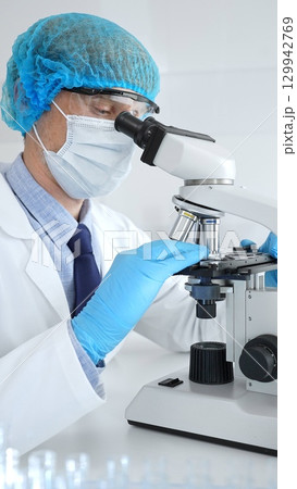 Senior male scientist wearing a protective mask, cap, and gloves, examining samples under a microscope in a modern laboratory. Medicine, healthcare and science concept Senior male scientist wearing a protective mask, cap, and gloves, examining samples under a microscope in a modern laboratory. Medicine, healthcare and science concept 129942769