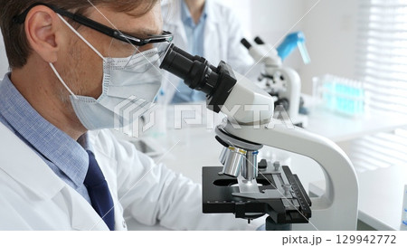 Medical researcher analyzing sample under microscope, wearing protective gear in sterile laboratory setting, performing scientific research. Medicine, healthcare and science concept Medical researcher analyzing sample under microscope, wearing protective gear in sterile laboratory setting, performing scientific research. Medicine, healthcare and science concept 129942772