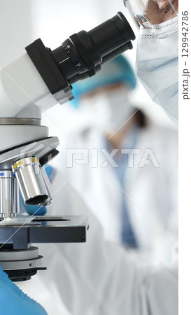 Close up of male researcher examining sample through microscope, wearing blue safety gear in modern laboratory setting. Medicine, healthcare and science concept Close up of male researcher examining sample through microscope, wearing blue safety gear in modern laboratory setting. Medicine, healthcare and science concept 129942786