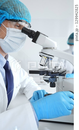 Senior male scientist wearing a protective mask, cap, and gloves, examining samples under a microscope in a modern laboratory. Medicine, healthcare and science concept 129942825