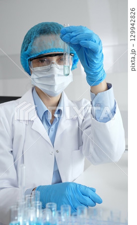 Close up of a scientist in ppe, holding a test tube with blue liquid while conducting research in a laboratory with a microscope. Medicine, health care and science concept 129942826