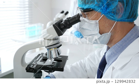 Male scientist in protective mask, cap and gloves studying samples under microscope in sterile laboratory. Concept of medicine, healthcare and science 129942832