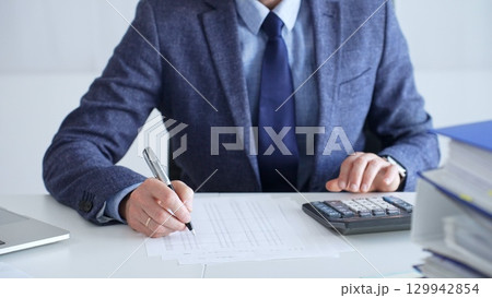 Businessman wearing suit and tie is using calculator and taking notes while working at desk in office, with laptop and stack of folders nearby, close up. Audit and taxes theme in business 129942854