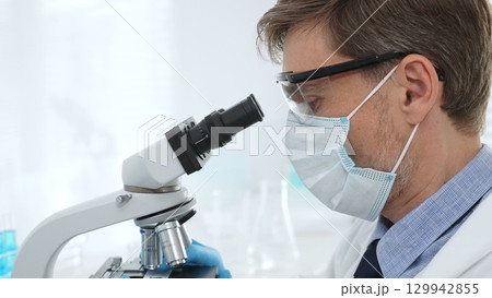 Scientist wearing mask and gloves using microscope doing scientific research in a laboratory with test tubes containing blue liquid. Medicine, healthcare and science concept Scientist wearing mask and gloves using microscope doing scientific research in a laboratory with test tubes containing blue liquid. Medicine, healthcare and science concept 129942855