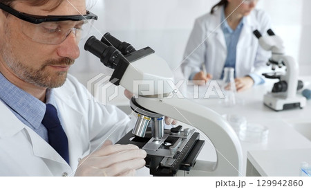 Researcher examining sample through microscope, wearing safety eyewear in sterile laboratory setting, surrounded by scientific glassware. Medicine, healthcare and science concept 129942860
